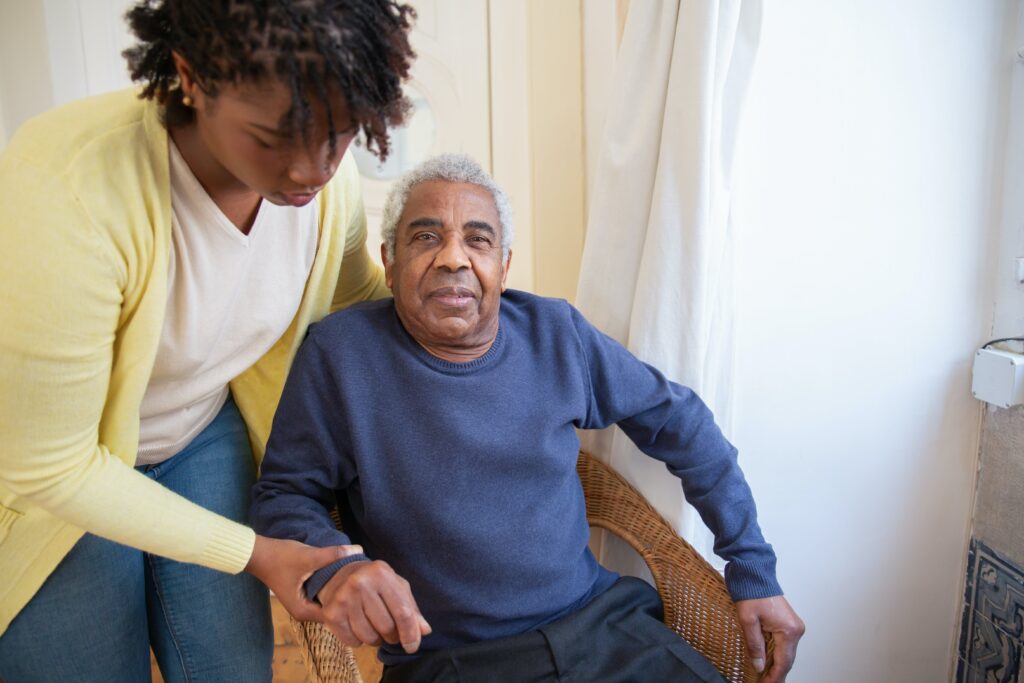 A young black woman helps an elderly black man out of a chair to find resources for black cancer patients together,