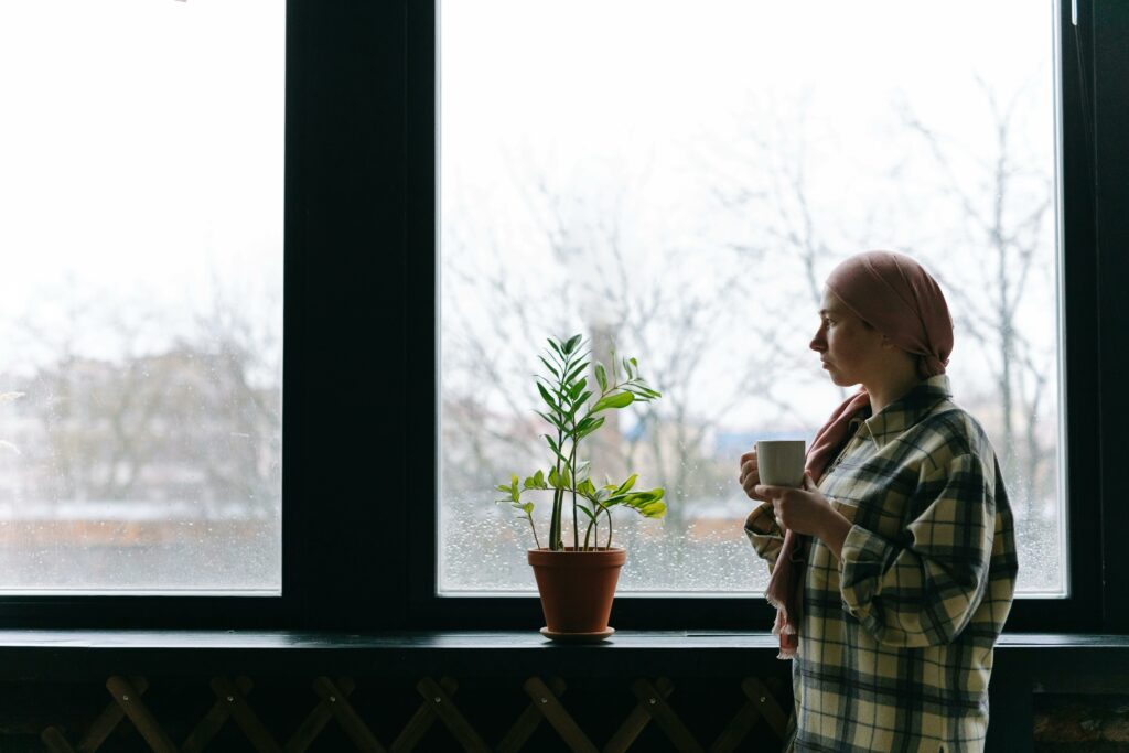 A cancer survivor looks out a large window while drinking tea from a mug.