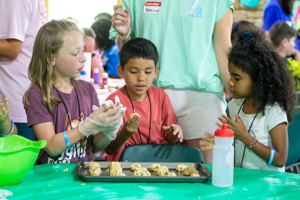 Two girls and one boy roll cookie dough into balls to place onto a baking sheet at CampCARE 2024.
