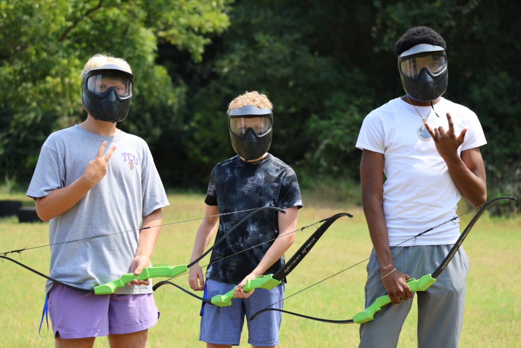 Three teenage boys wearing black helmets pose while holding archery tag bows at Teen Retreat 2024.