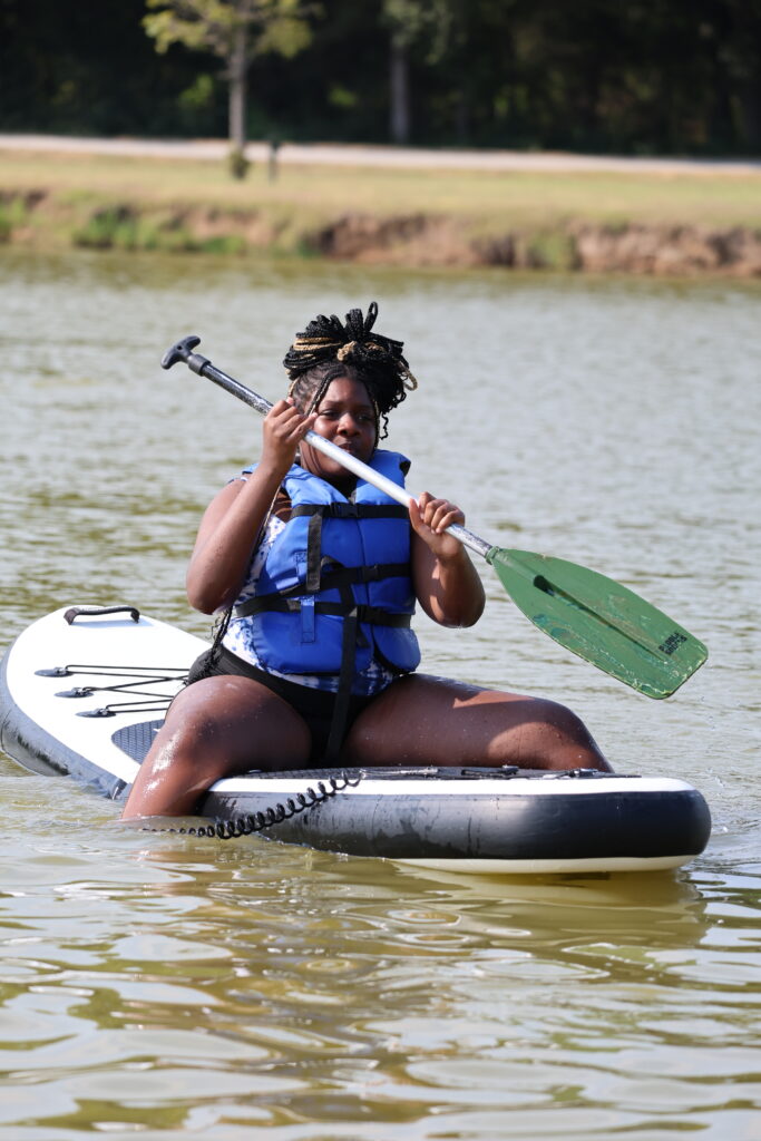 A teenage girl sits on a paddle board in the middle of a lake at Teen Retreat 2024.