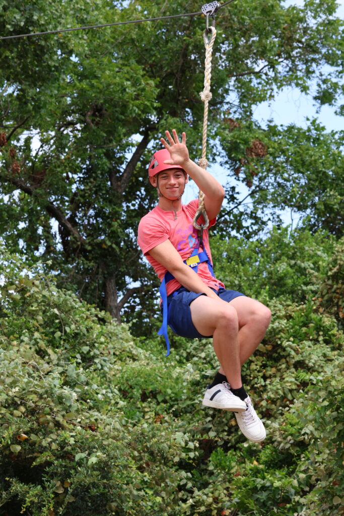 A teenage boy waves as he zips past on a zipline at Teen Retreat 2024.